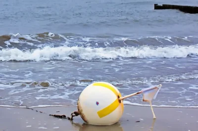 Gelb-weiße Boje mit Flagge am nassen Sandstrand vor brechenden Wellen der Ostsee im Detail.