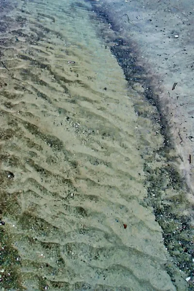 Klares, flaches Meerwasser spült über sandige Rippelmarken (Sandwellen) am Küstenstrand im Detail.