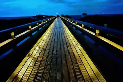 Beleuchtete Seebrücke aus Holz bei Heiligenhafen bei Nacht mit Spiegelung auf feuchten Planken unter blauem Himmel.